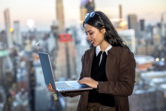 Young businesswoman stands on a manhattan rooftop, smiling and working on a laptop with the new