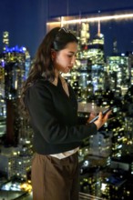 Young businesswoman standing by a window, engrossed in her smartphone, with the illuminated