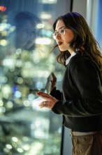 Young businesswoman standing by a skyscraper window, looking thoughtfully at the vibrant night city