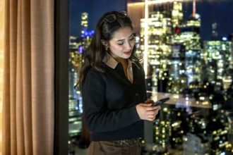 Young businesswoman standing by a window, focused on her smartphone while managing business