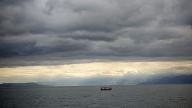 Small boat on calm sea under cloudy sky surrounded by mountains in the background, Lake Ohrid,