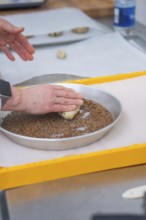Hands press dough into a round dish filled with seeds on a yellow tray, bake rolls, Haselstaller