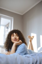 A woman is lying on her stomach on a bed with her legs in the air. She looks thoughtful while