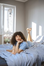 A young woman is lying on her stomach on a bed covered with blue sheet. She rests her chin on her