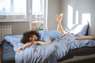 A young woman lies on her stomach on a bed, wearing blue pajamas. Sunlight comes through the window