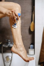 Unrecognizable woman put her leg on the edge of a bathtub and using a razor to shave her leg.