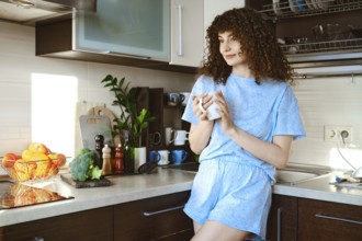 Smiling woman stands in a kitchen, sipping coffee from a mug. Fresh fruits and vegetables are on