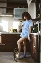 A woman is in the kitchen wearing a light blue outfit. She holds a drink in one hand while her cat