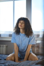 A woman sits on a bed in front of a large window. The sun shines in as she looks at the camera. She