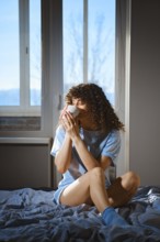 A woman sits on a bed with a blue blanket, holding a white cup. Bright winter sunlight comes