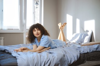A woman with curly hair lays on a bed with light blue sheets. She looks to the ceiling while