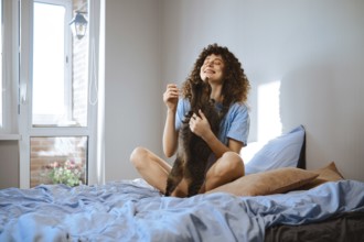 A woman sits on her bed with pillows and blue sheets. She smiles while holding and playing with a