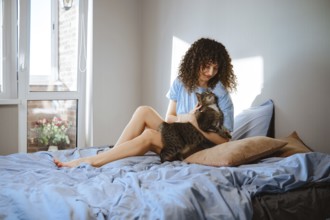 A woman sits on a bed and plays with a cat in a bright room. Sunlight shines through the window,