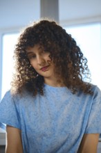 A woman with curly hair sits by a window. She wears a blue shirt and looks down with a thoughtful