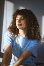 A woman with curly hair sits in a well-lit room. Sunlight streams through the window, creating warm