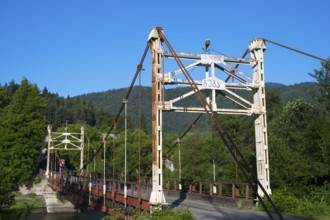 Old suspension bridge stretches across a river surrounded by green landscape, suspension bridge