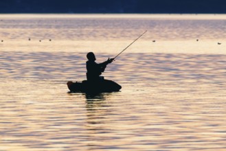 Angler sport fisherman in his belly boat in orange-red lake at sunset, silhouette in calm water,