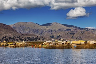 Scene with traditional huts on a lake against a mountain backdrop under blue skies and clouds, The