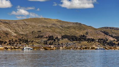 A calm lake in front of a mountainous landscape with boats and a village on the shore under a blue