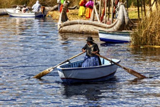 A woman in traditional dress rows a boat across a calm lake, the floating reed islands of the Uros