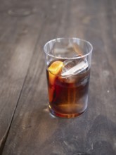Glass of dark alcoholic cocktail with ice and orange zest on a rustic wooden table
