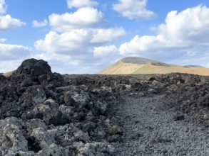 Hiking trail through lava rock to Caldera Blanca volcano against blue sky with white clouds, Mancha