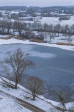 Small lake surrounded by snow-covered park and village in the background, icy surface, Empfingen,