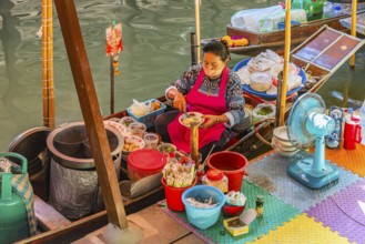 Elderly woman wearing mask offers food on her wooden boat, Damneon Saduak floating markets channel,