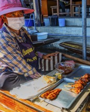 Elderly woman wearing mask sells barbecue skewers on her wooden boat on the Damneon Saduak floating