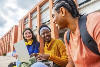 Diverse group of university students collaborating and smiling around a laptop outdoors on campus,