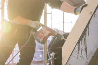 Worker with drill in sunlight working on wood on construction site, carpentry construction, roof
