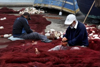 Fishermen repair nets in harbor surrounded by blue boats and tools, Essaouira, Marrakesh-Safi,