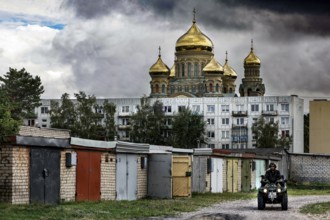 In front of an imposing church, golden domes rise above prefabricated buildings and garages,
