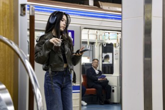 Woman standing in Osaka subway looking at her smartphone while a man sits in the background, Osaka,