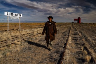 Man wearing cowboy clothes on abandoned tracks in front of station sign and water tower, Oodnadatta
