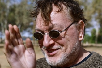 Smiling man wearing sunglasses against a tree-filled backdrop in a relaxed atmosphere, William