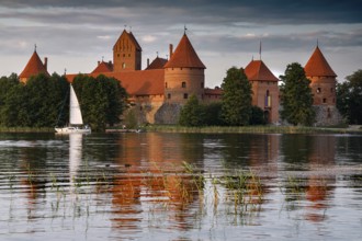 Trakai castle with red roof on a lake surrounded by trees and a sailboat, Trakai, Lithuania