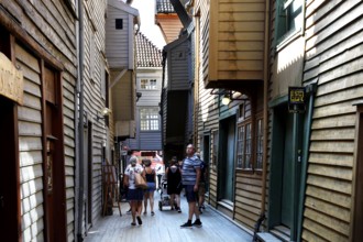 Narrow wooden house alley in Tyskebryggen with passers-by in sunshine, mountains, Hordaland, Norway
