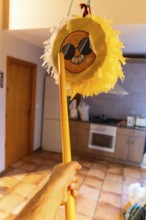 Hand holding yellow stick poised to strike a hanging emoji pinata in a home interior, playful