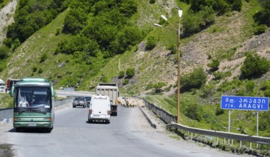 A road with vehicles in a mountainous landscape, green hills and a sign with a river name, bridge