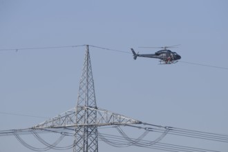 A helicopter carries out technical work on power lines next to a mast under a blue sky, an employee