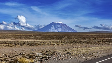 Volcano in a wide desert landscape under blue sky with clouds, snow-capped summit, The Andean