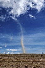 A narrow tornado crosses a dry plain, the sky is clear with scattered clouds and the mountains are