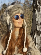 Slender pretty woman with fur hat and pompoms, red hair and warm winter colors between birch forest