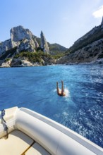 Young woman jumping from a boat into the water, picturesque rocky coast, cliffs with L'Aguglia rock
