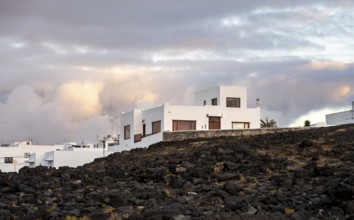 Black coast of volcanic rocks behind typical white houses of the village of La Santa, at sunset,