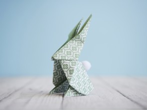 Green origami bunny standing on a white wooden table with a light blue background, representing