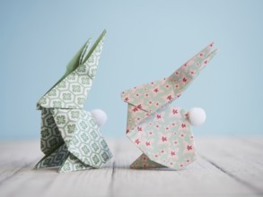 Two colorful origami bunnies standing on a white wooden table with a light blue background, perfect