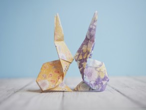 Two colorful origami bunnies are facing each other, touching noses, on a wooden surface with a