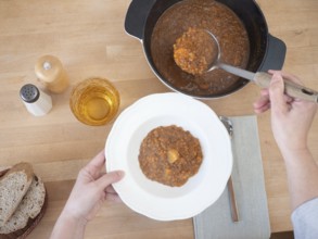 Hands serving lentil soup with potatoes in a white plate on a wooden table, healthy eating concept